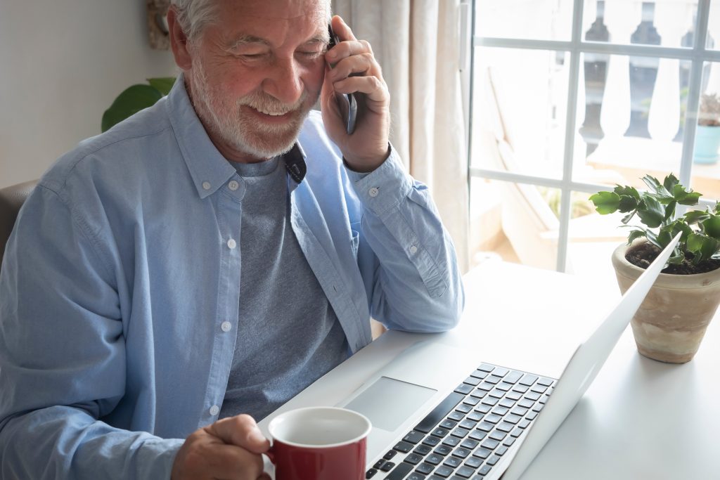 Mature man talking on the telephone while looking at his laptop and drinking a mug of coffee