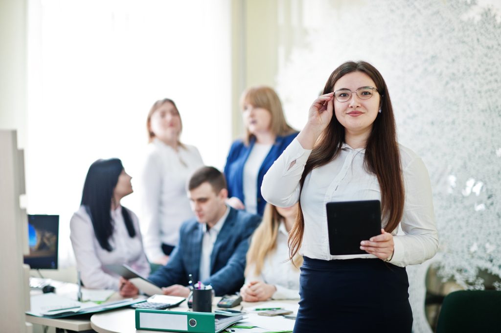 Young caucasian woman wearing eyeglasses holding a tablet, standing against a backdrop of a group of office workers.